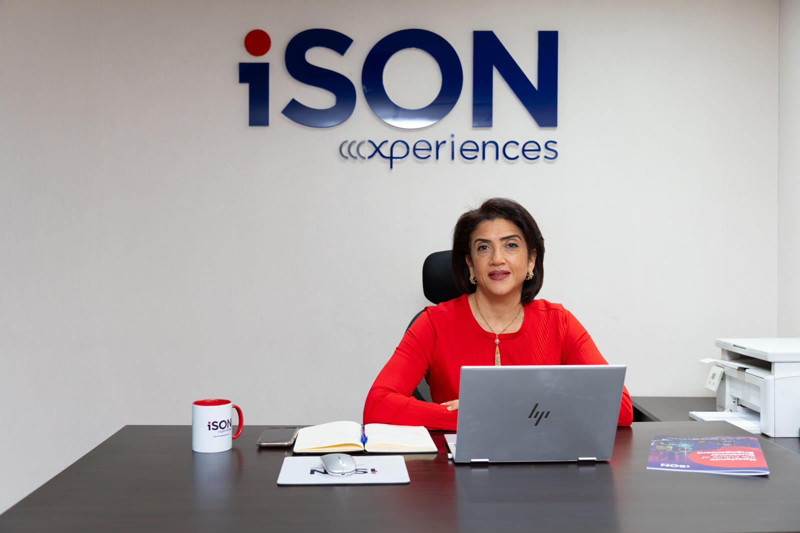 Amani Rabei, Chief Growth Officer, iSON Xperiences, Egypt, Offshore BPO Confidence Index 2023: Woman in a red top seated at a desk with a laptop beneath an iSON Xperiences logo on the wall