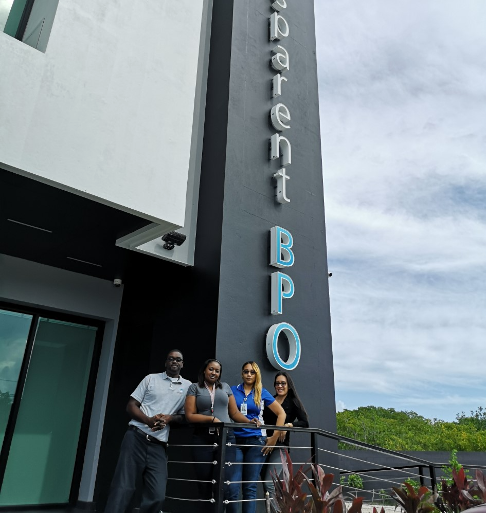 Transparent BPO team in Belize, Offshore BPO Confidence Index 2023: Four employees standing on a balcony beside a tall Transparent BPO sign on the building front