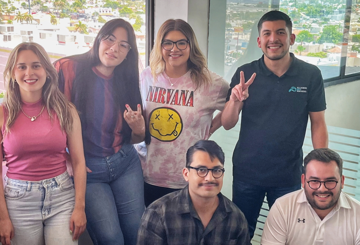 Alliance BPO Services team in Mexico, Offshore BPO Confidence Index 2024: Six smiling people posing by floor-to-ceiling windows in an office, four standing and two crouching in front