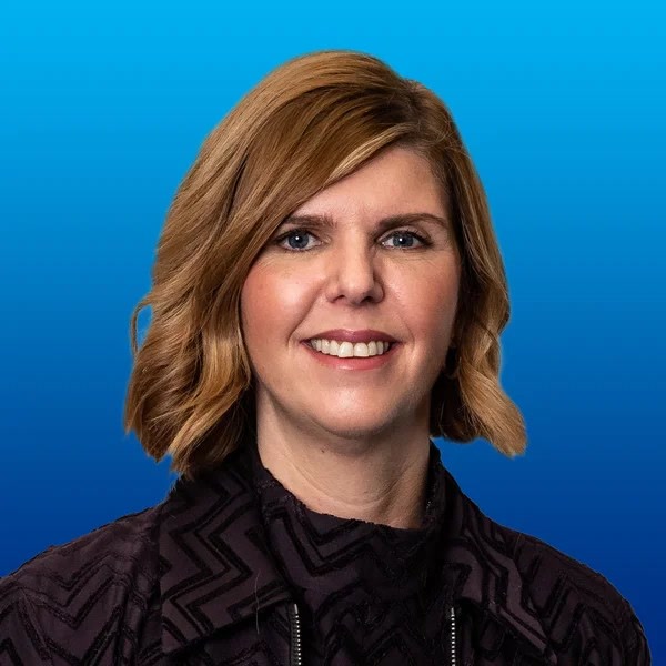 Carrie Ramskill, COO, UK and South Africa, HGS South Africa, Offshore BPO Confidence Index 2024: Smiling woman with shoulder-length auburn wavy hair and a dark patterned top against a bright blue background