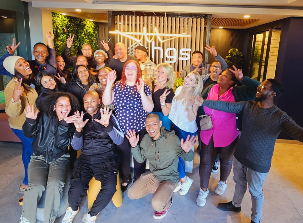HGS South Africa team, Offshore BPO Confidence Index 2024: About twenty employees posing and making hand gestures in front of a glowing hgs sign