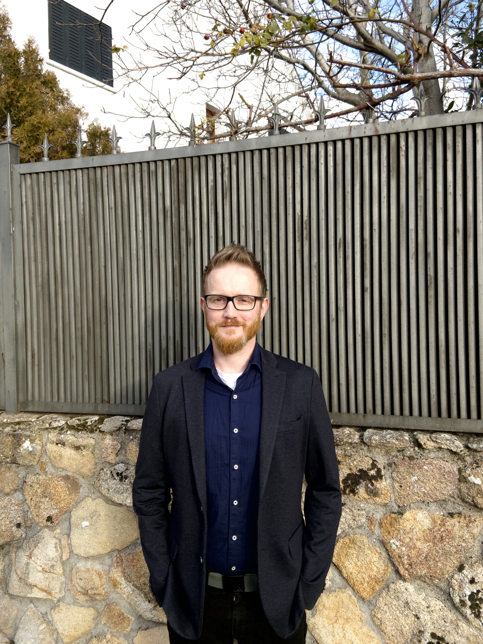 Matt Kendall, Founder, Off Mute Media, author of the Offshore BPO Confidence Index 2024: Man with glasses and a dark blazer, hands in pockets, before a stone wall and metal fence