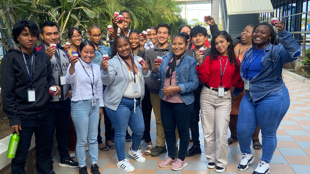 OneSource team in Belize, Offshore BPO Confidence Index 2024: About fifteen young people posing outdoors under palm trees, several holding small cupcakes up for the camera