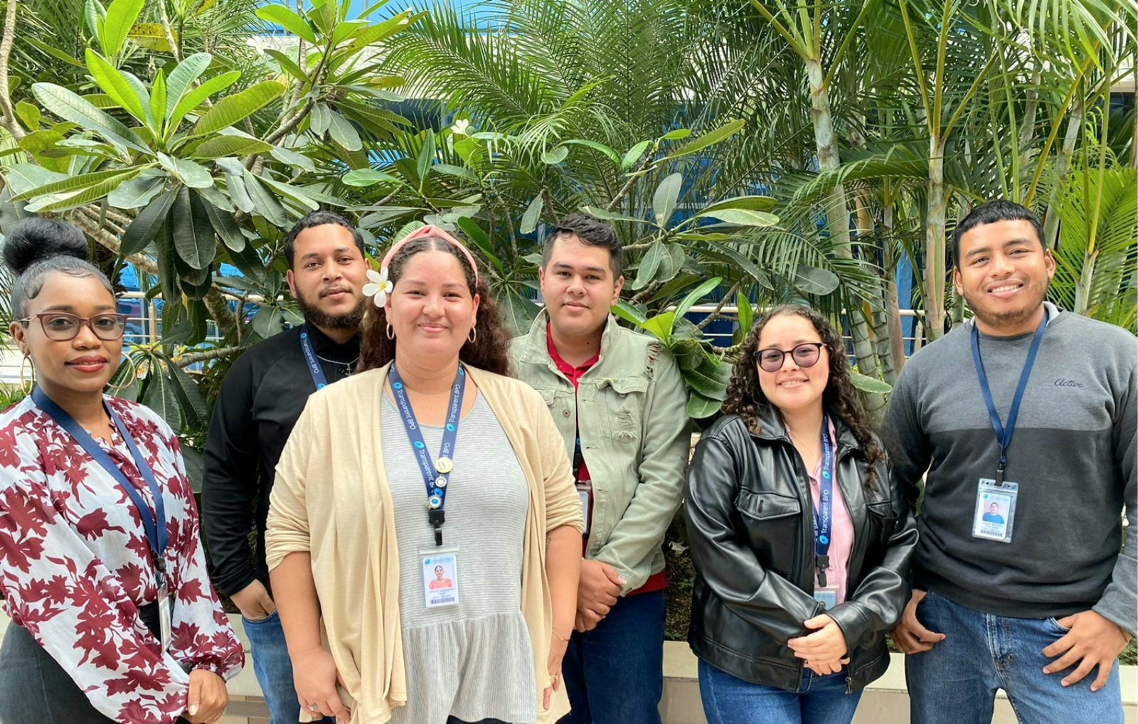 OneSource team in Belize, Offshore BPO Confidence Index 2024: Six employees with lanyards and ID badges posing together outdoors among tropical plants and palm trees