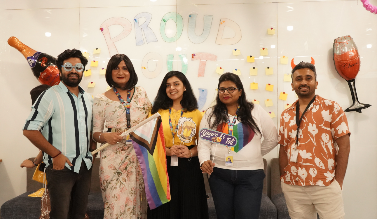 TaskUs India team, Offshore BPO Confidence Index 2024: Five people posing in an office in front of a PROUD OUT wall, one holding a Pride flag