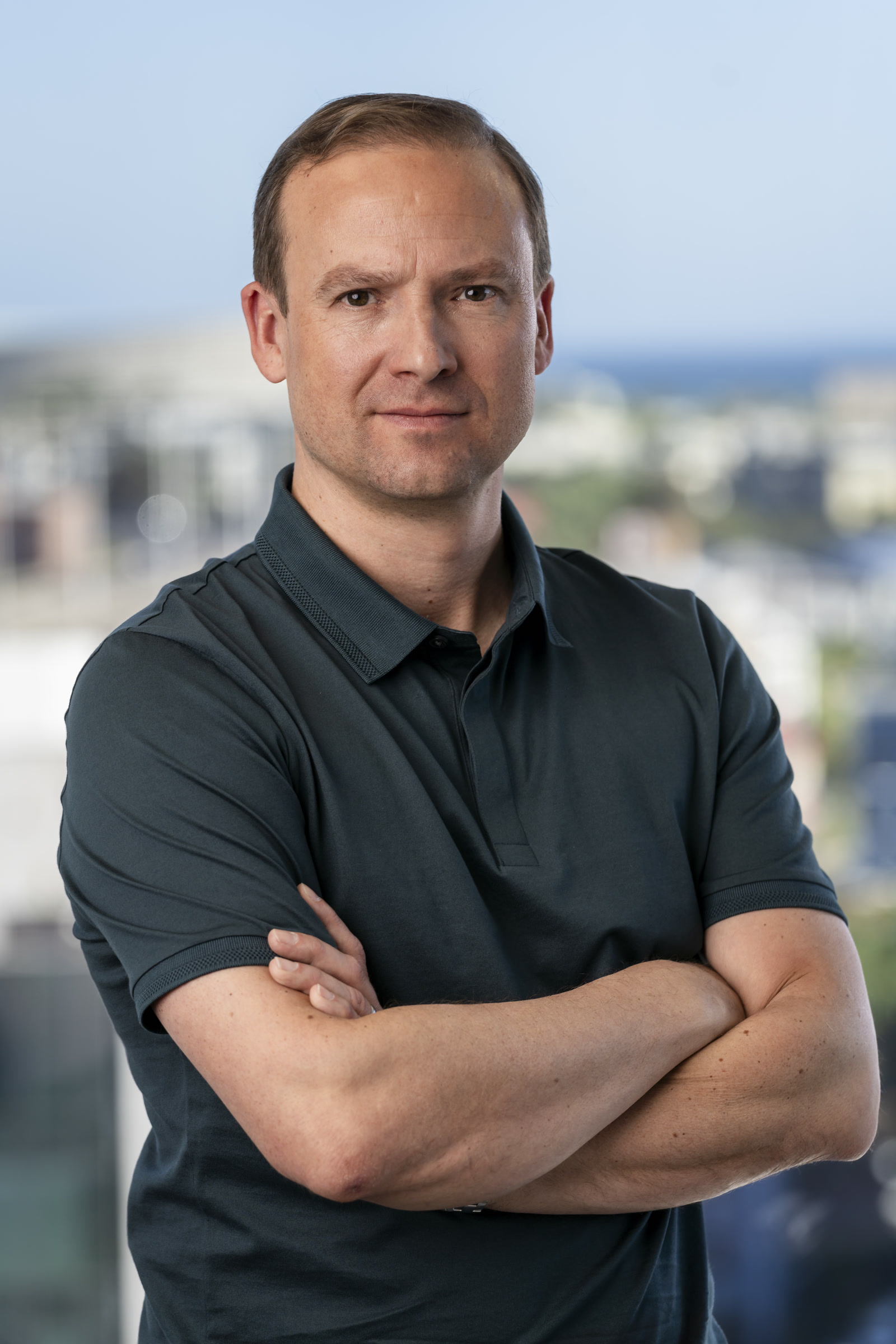 Alastair Brass, Founder & CEO, Amplify5, South Africa, Offshore CX Confidence Index 2025: Man with short hair in a dark gray polo shirt, arms crossed, before a blurred city and sea