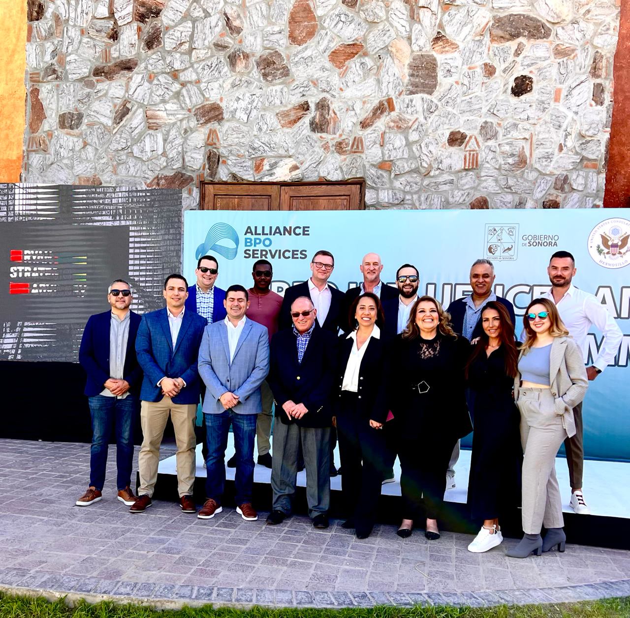 Alliance BPO Services team in Mexico, Offshore CX Confidence Index 2025: About twenty people in business attire posing in front of a stone wall and a branded banner