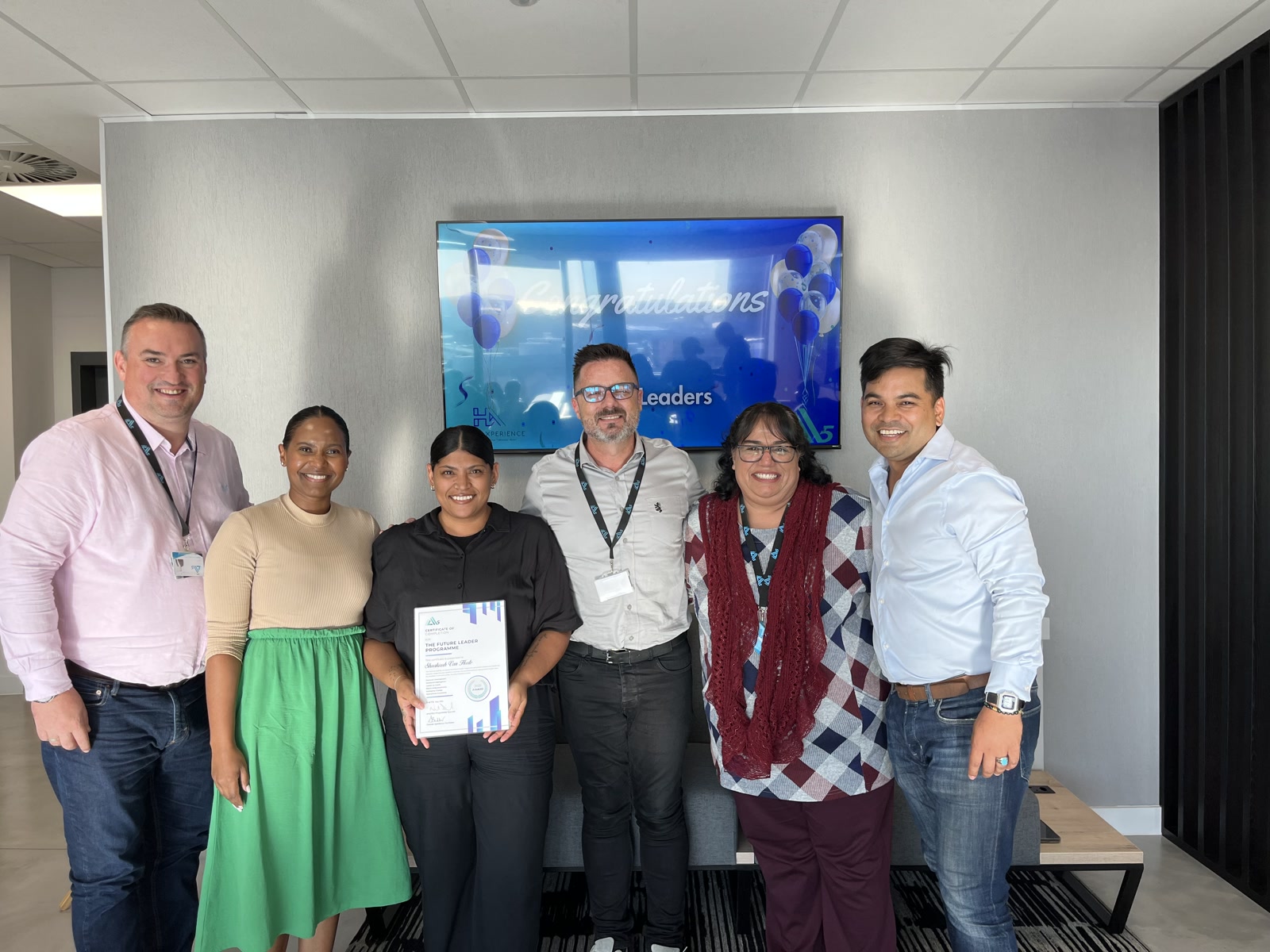 Amplify5 team in South Africa, Offshore CX Confidence Index 2025: Six people smiling in an office, one holding a certificate, with a screen reading Congratulations behind