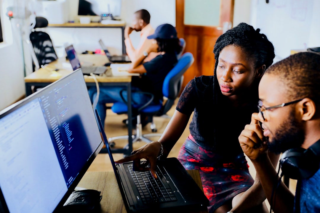 Contax360 BPO Solutions team in Jamaica, Offshore CX Confidence Index 2025: Two coworkers study a laptop and a monitor showing code while colleagues work at desks behind them
