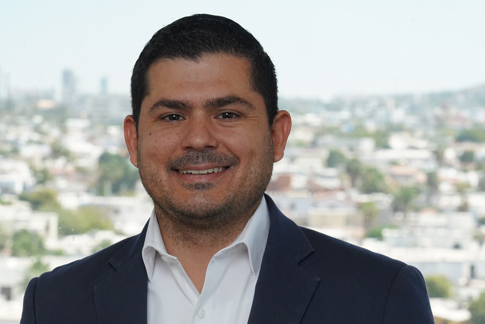 Juan Caire, Chief Executive Officer, Alliance BPO Services, Mexico, Offshore CX Confidence Index 2025: Smiling man with short dark hair in a navy blazer and white open-collar shirt before a blurred city