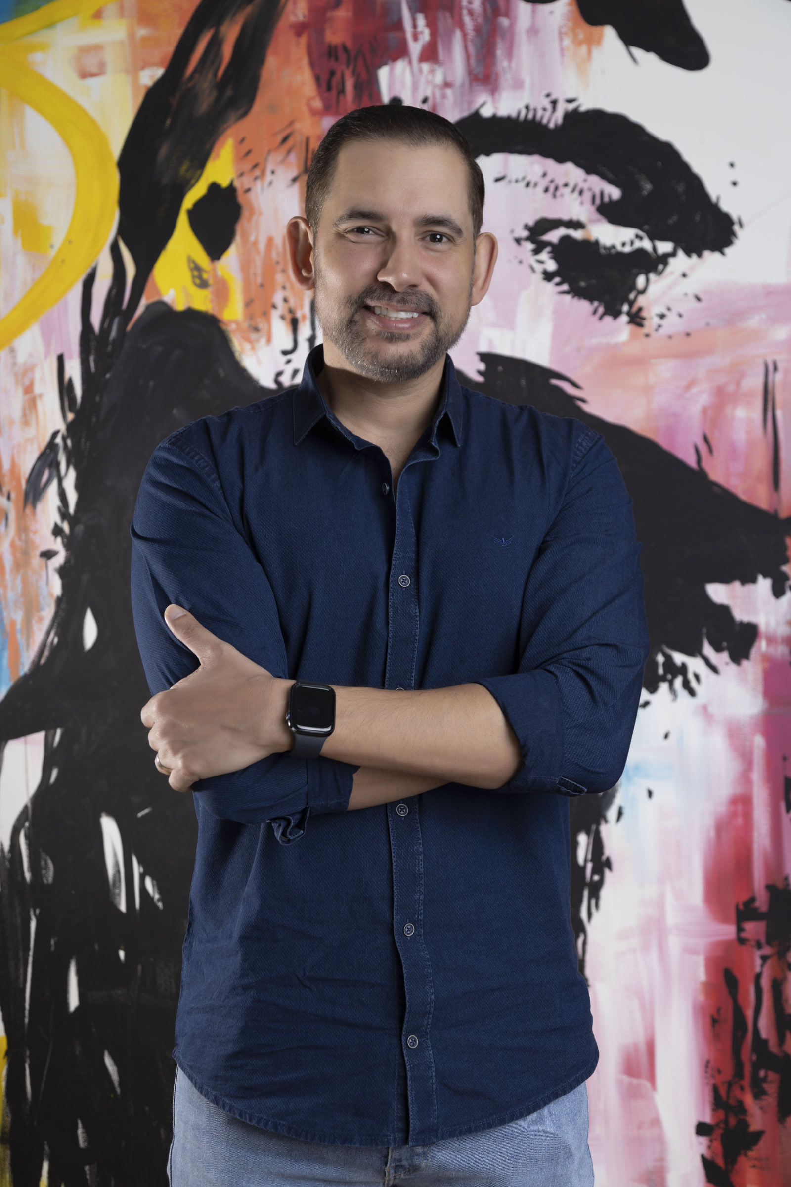 Luis Barreto, Division Vice President LATAM, TaskUs, Colombia, Offshore CX Confidence Index 2025: Smiling man with arms crossed in a navy shirt, standing before a black, pink, and orange painting