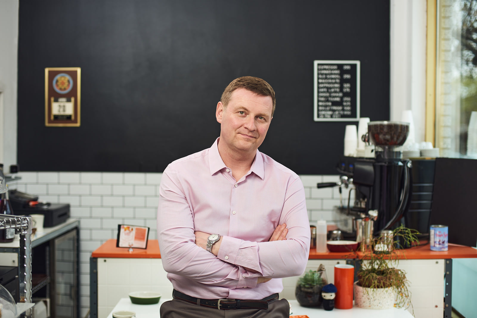 Peter Ryan, Principal, Ryan Strategic Advisory, author of the Offshore CX Confidence Index 2025: Man with folded arms and slight smile in pink shirt, in front of a coffee bar chalkboard