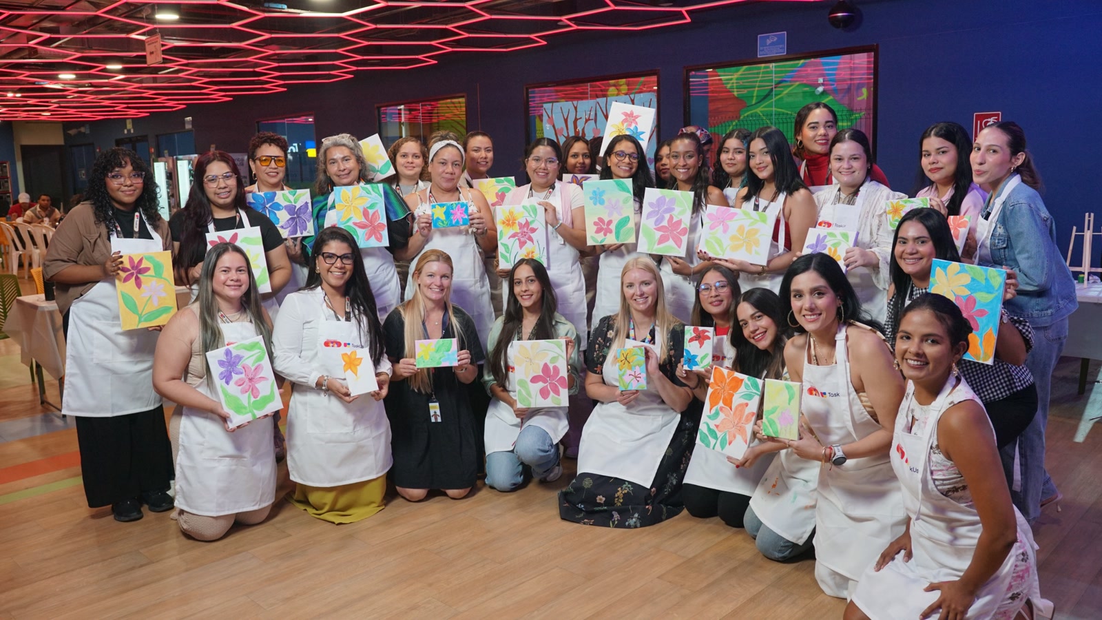TaskUs team in Colombia, Offshore CX Confidence Index 2025: About thirty employees in white aprons holding painted flower canvases indoors under red ceiling lights