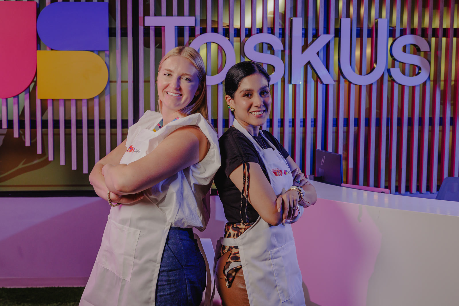 TaskUs team in Colombia, Offshore CX Confidence Index 2025: Two smiling women in white aprons stand back to back before a pink and purple lettered wall