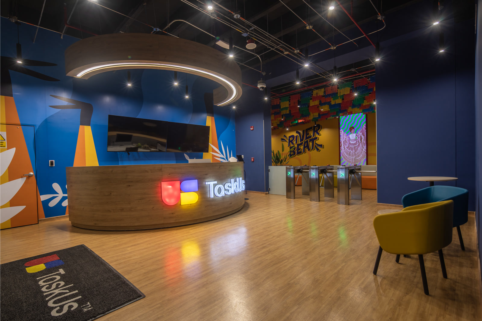 TaskUs team in Colombia, Offshore CX Confidence Index 2025: Empty office lobby with curved wood reception desk, blue mural walls, and a lit TaskUs sign