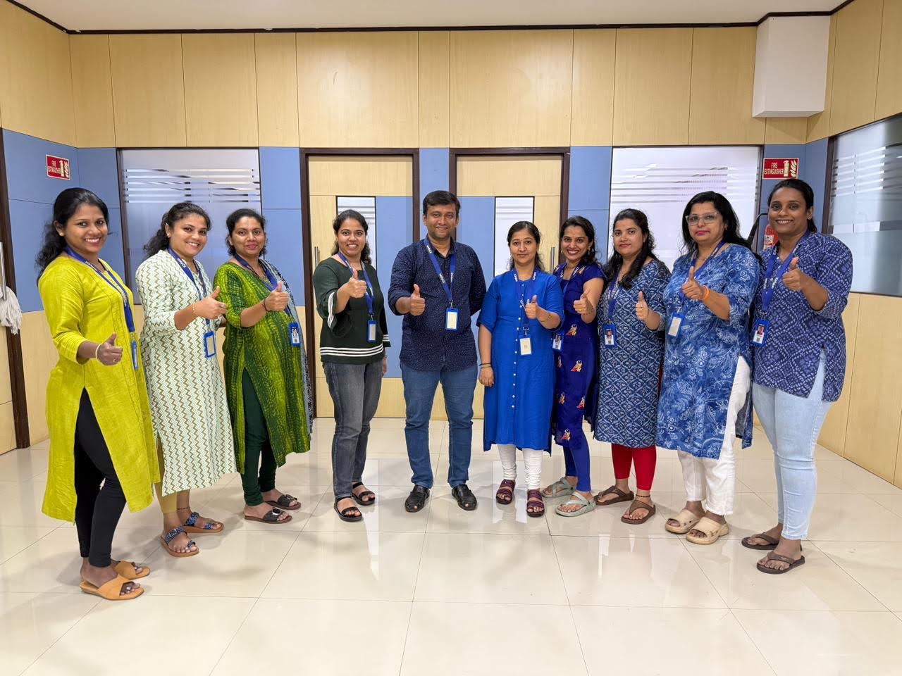 UnifyCX team in India, Offshore CX Confidence Index 2025: Ten employees in long tunics give thumbs up in a bright office lobby with wood-paneled walls