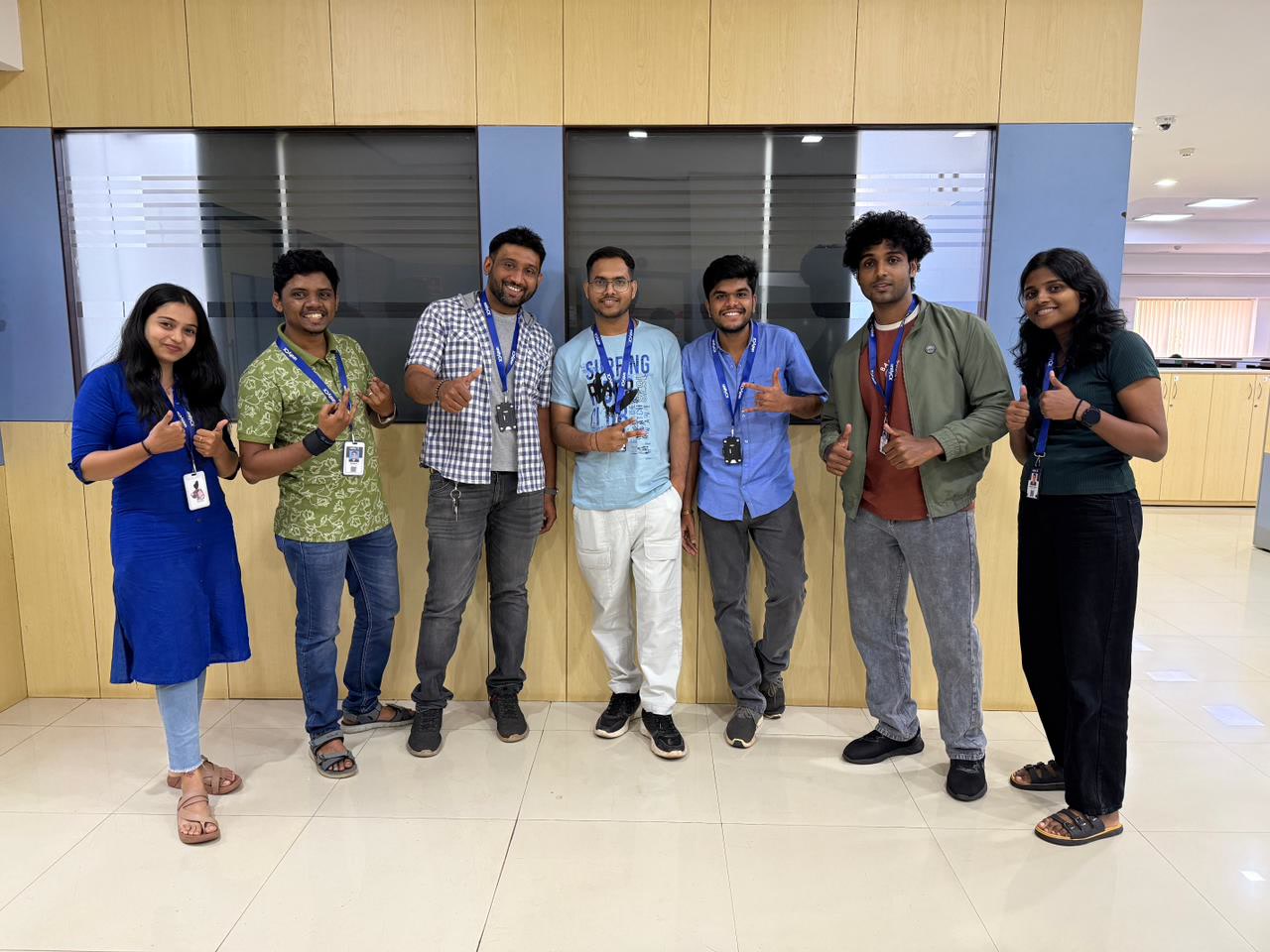 UnifyCX team in India, Offshore CX Confidence Index 2025: Seven young employees giving thumbs-up gestures in front of wood-paneled office cabinets, wearing blue lanyards